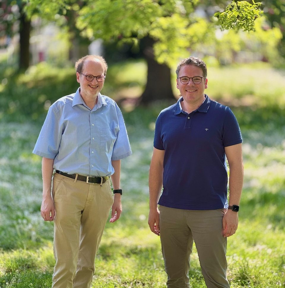 Zwei Männer stehen lachend in einem sonnigen Park mit einer grünen Wiese im Hintergrund Zwei Männer stehen lachend in einem sonnigen Park mit grüner Wiese im Hintergrund.